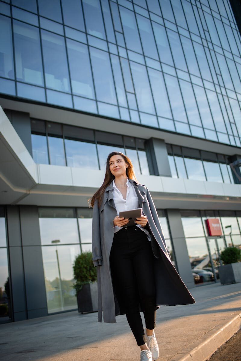 A professional businesswoman in a gray coat walks near a modern glass building holding a tablet and smiling.