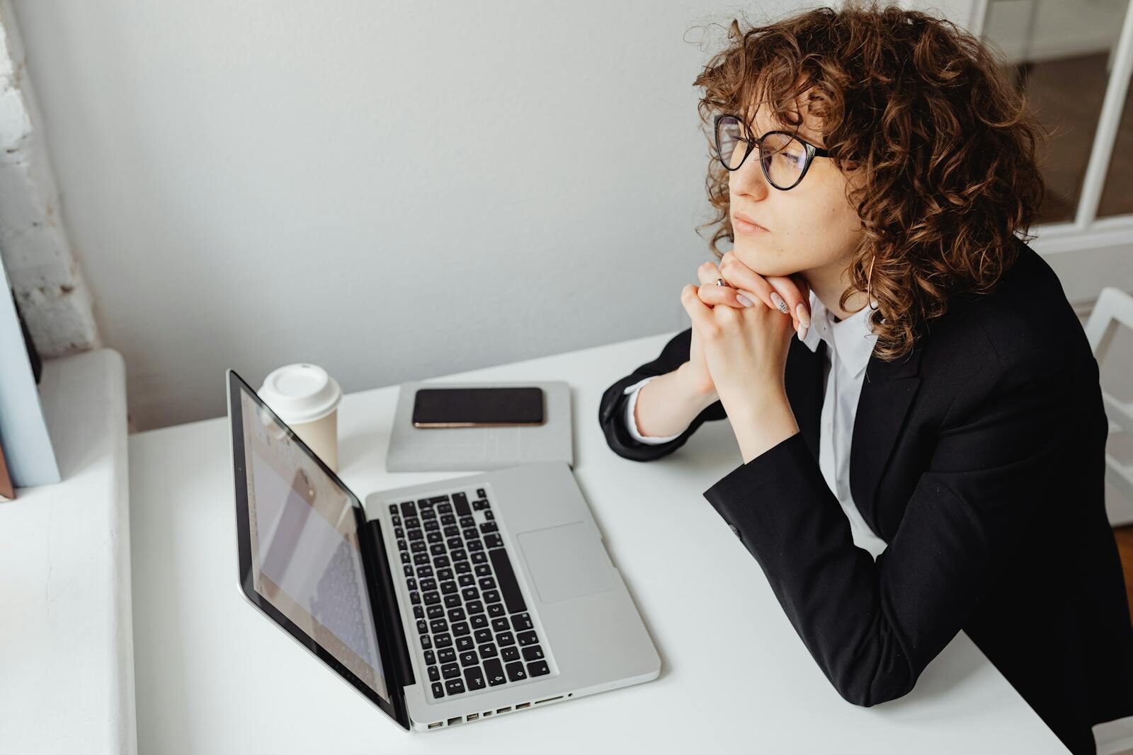 A pensive woman in business attire sits at a desk with a laptop, contemplating work.
