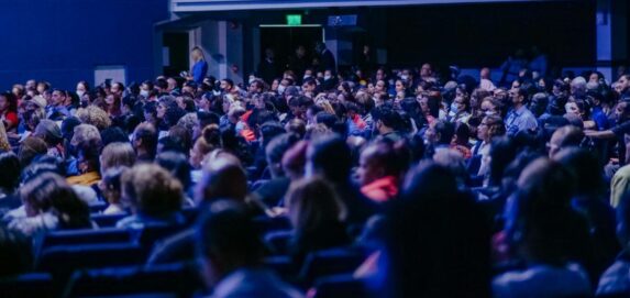 A crowded conference hall with diverse audience attentively listening to a speaker.