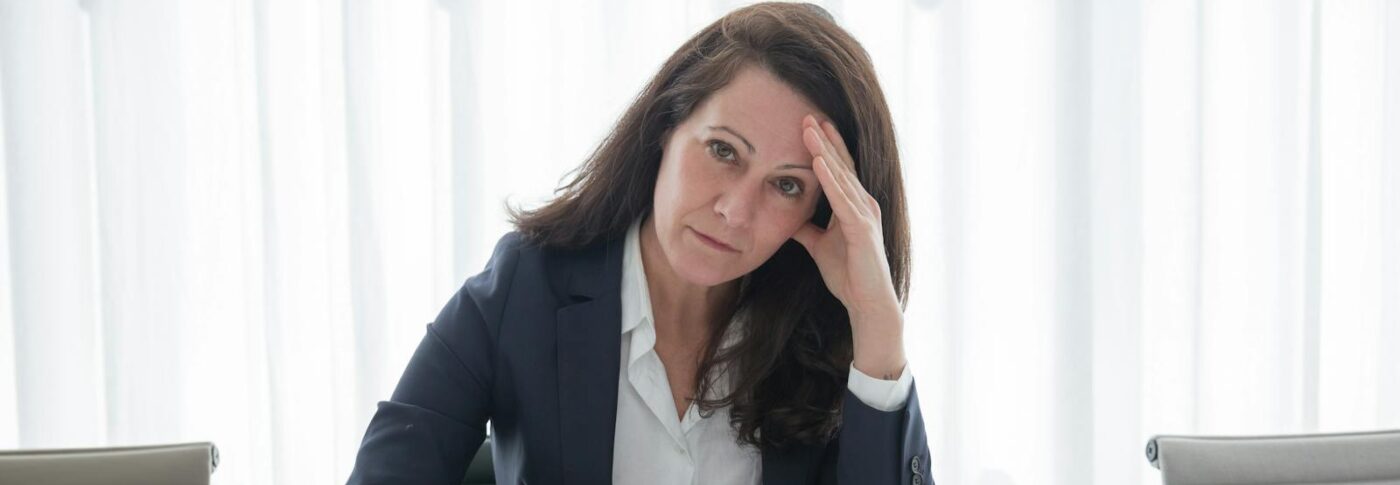 A stressed businesswoman in corporate attire sits at a desk with papers, looking overwhelmed.
